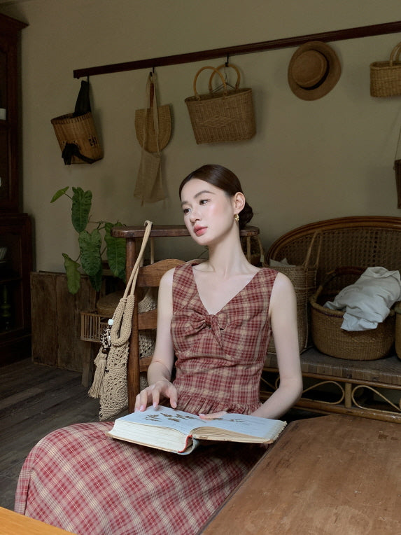 Reading a floral book in a rustic room, a woman wears a vintage check dress with ribbon accents, evoking a peaceful and romantic vibe.