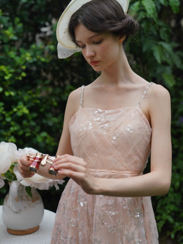 Model delicately holds opera glasses beside a flower table, wearing a sequin-embroidered blush dress with pearl straps in a vintage fairycore mood.