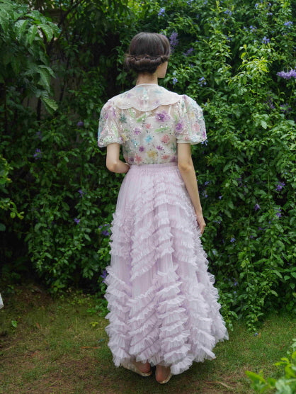 Model stands in a lush garden wearing a pastel lavender layered skirt and vintage-style floral blouse, evoking a soft fairycore aesthetic charm.