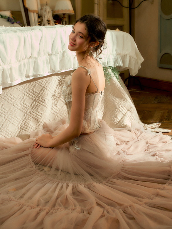 Smiling softly, the model sits on the floor in a voluminous blush tulle skirt and lace-trimmed corset, glowing in dreamy fairycore light.