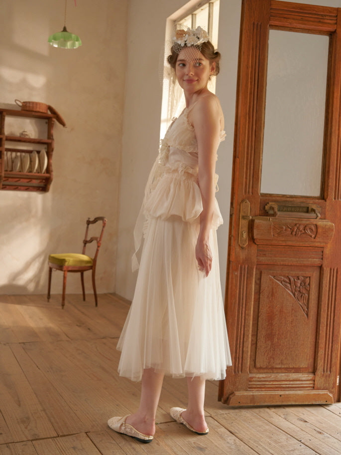 Side view of a model in a fairytale-inspired ivory layered dress with a floral waist accent and soft tulle skirt, walking on wooden flooring.