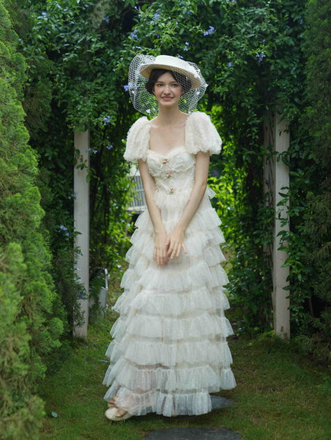 Model smiles softly in a ruffled ivory dress with puff sleeves and floral bows, framed by greenery for a whimsical princesscore garden look.