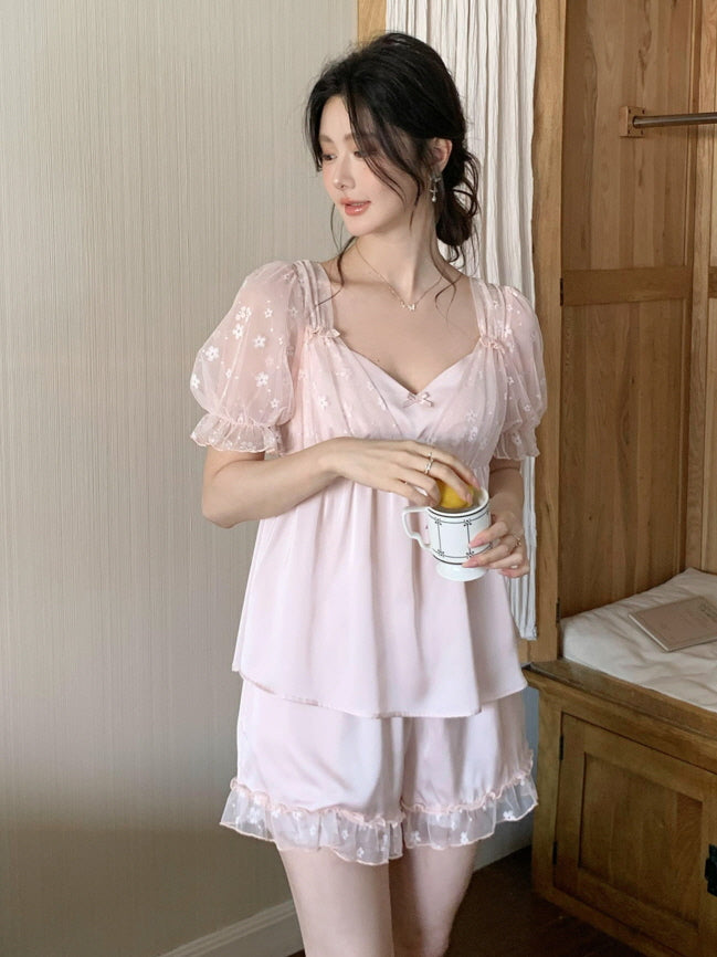 A woman in a blush pink coquette loungewear set with embroidered puff sleeves enjoys a quiet morning with coffee and bread in a cozy cottage bedroom.