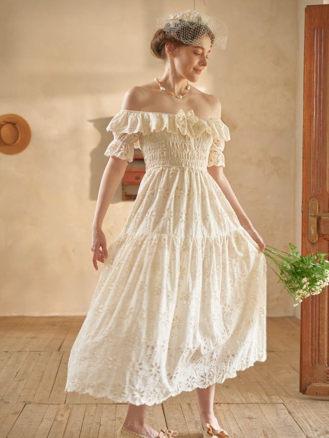 A woman twirls slightly in a cream lace dress, her hand brushing the skirt while holding fresh flowers, capturing a soft fairytale moment.