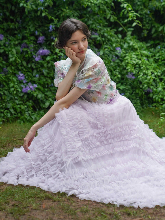 A model sits gracefully in a blooming garden, wearing a pastel lilac ruffle skirt and a floral embroidered sheer blouse for a fairycore mood.