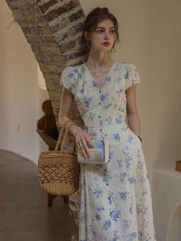 A woman in a cottagecore-style floral dress with puff sleeves and lace detailing stands against a brick archway, holding a woven bag and a book, creating a dreamy, romantic vibe.