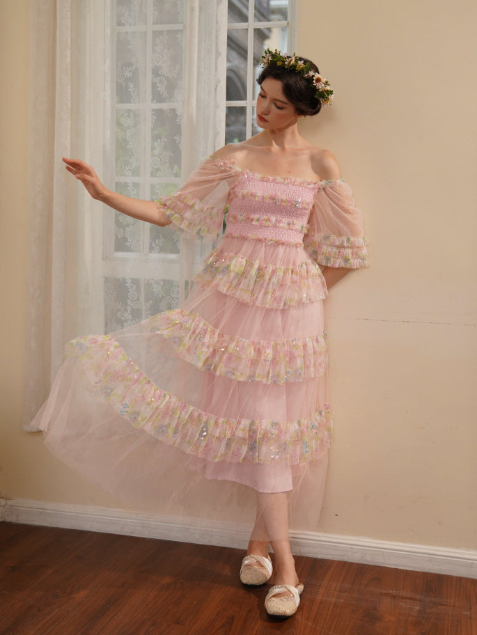 A woman in a pink floral ruffle dress gently lifts the sheer hem with one hand, striking a graceful pose in a dreamy fairycore room.