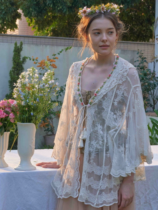 Wearing a vintage lace robe over a corset top, the model poses beside a flower-covered table in a soft cottagecore setting.