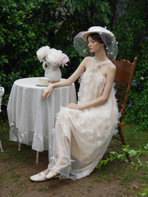 Sitting beside a floral tea setting, the model wears a beige tulle dress with 3D flowers and a wide-brim hat, evoking a peaceful cottagecore mood.
