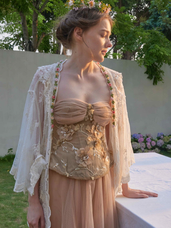 The model stands in sunset light wearing a floral embroidered corset and sheer robe, styled for a dreamy fairycore garden look.