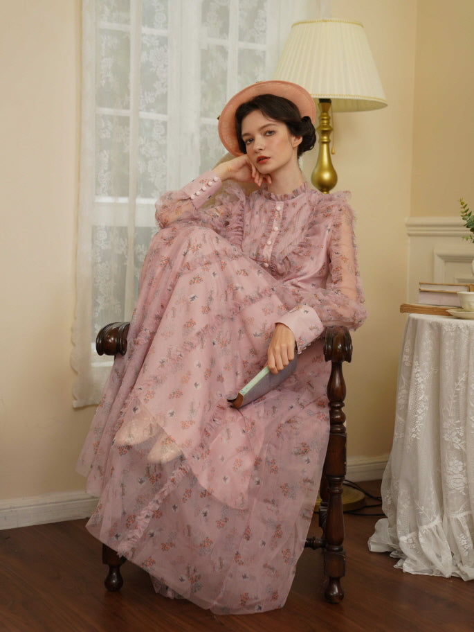 The model sits thoughtfully in a vintage chair, wearing a sheer pink ruffle dress with floral details and soft coquette charm.