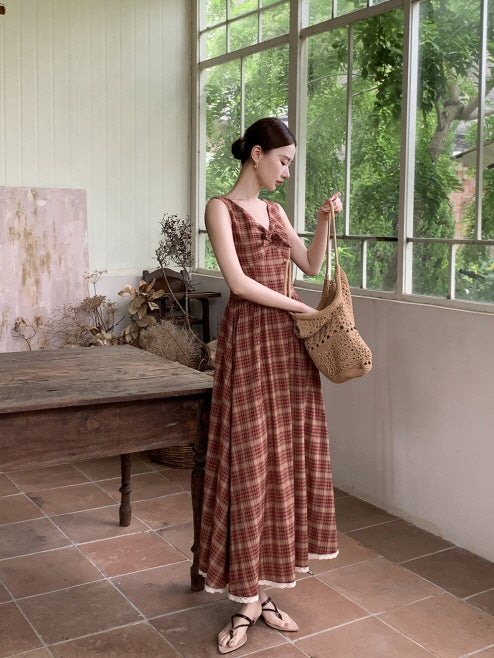 Holding a woven shoulder bag, a woman in a checkered country-style dress stands near a wooden table in a sunlit rustic room.