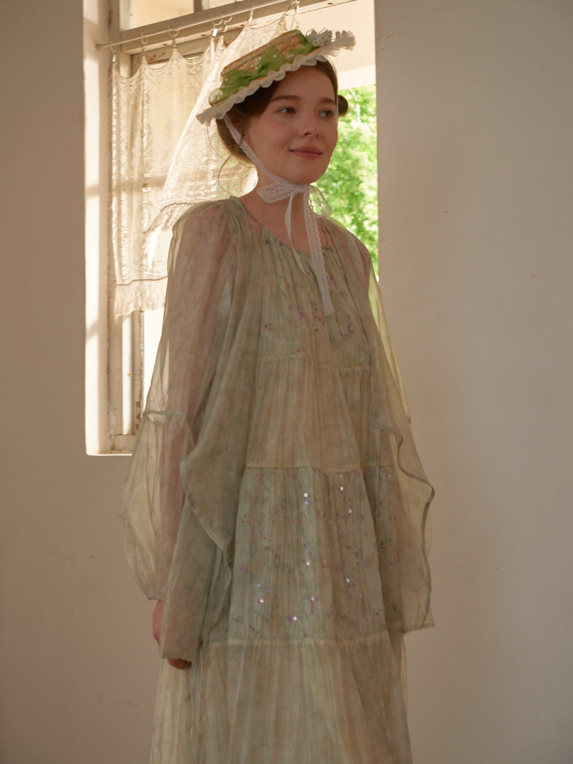 Smiling by the window, the woman wears a shimmering floral tiered dress with a translucent overlay, embodying soft fairycore elegance.
