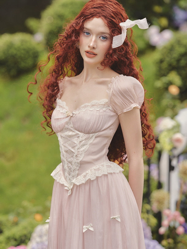 A young woman with long red curls wears a vintage-inspired puff-sleeve fairytale dress with lace and ribbon details, standing in a blooming garden.