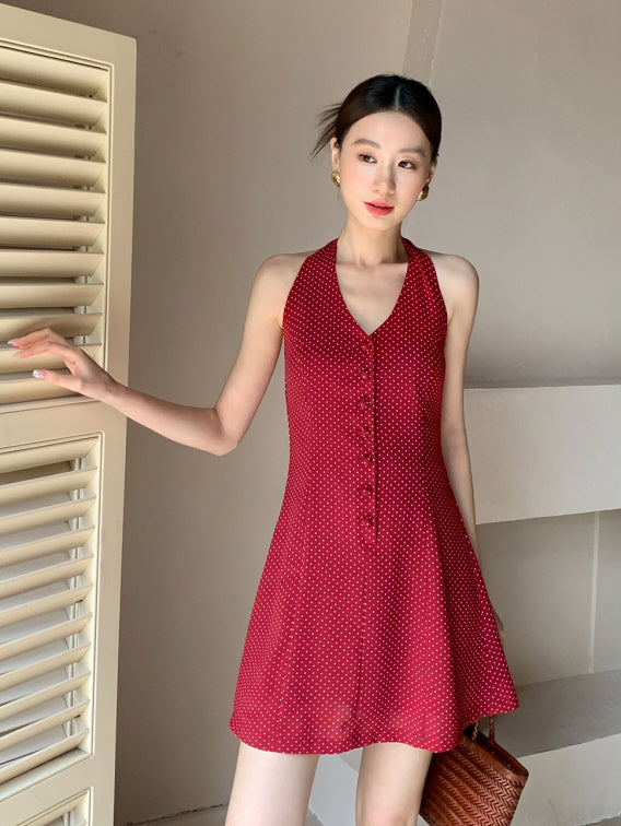 Woman gazes softly while wearing a red halter mini dress with dainty polka dots, styled with gold earrings and a woven bag for a coquette summer look.