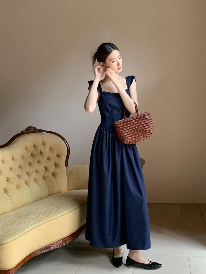 A woman in a navy fairytale-inspired dress adjusts her earring beside a vintage sofa, holding a woven bag with soft vintage charm.