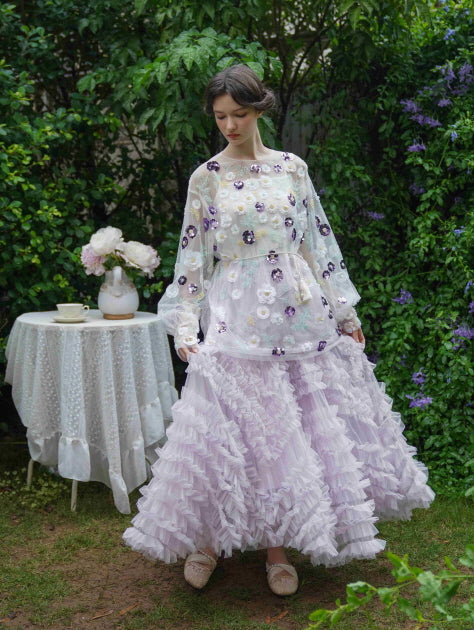 A romantic fairytale outfit featuring a lilac tiered tulle skirt and flower-appliqué top, captured as the model strolls softly through a lush green garden.