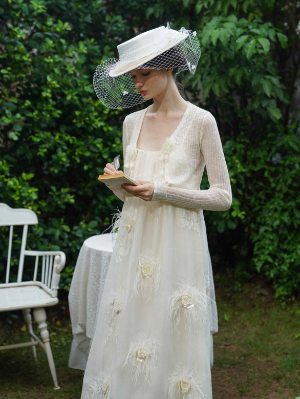 Model writes in a notebook wearing a cream floral feather dress and soft cardigan, styled in a vintage garden for a romantic fairycore look.