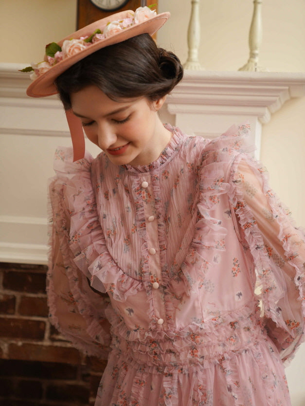 Smiling gently, the model in a pink fairycore dress with frilled buttons and floral print exudes romantic vintage elegance by the fireplace.