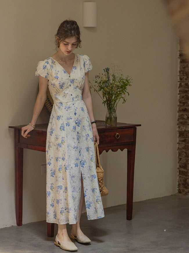 A woman gently touches a wooden table while wearing a cream floral dress adorned with blue blooms. The dress has a front slit and delicate cap sleeves, creating a romantic, countryside-inspired look.