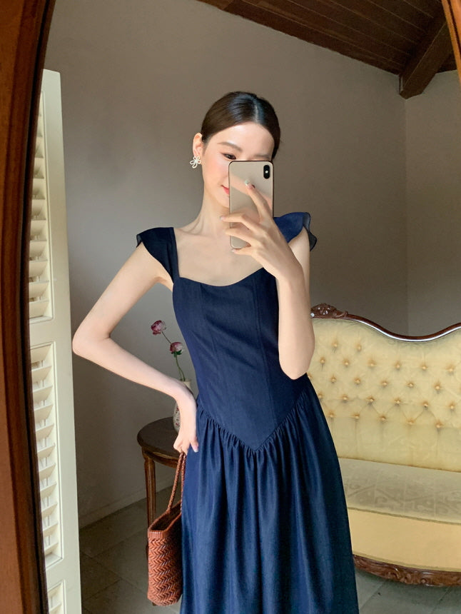Taking a mirror selfie in a navy feminine summer  dress, the woman stands with a relaxed pose in front of a tufted vintage sofa, radiating quiet elegance.