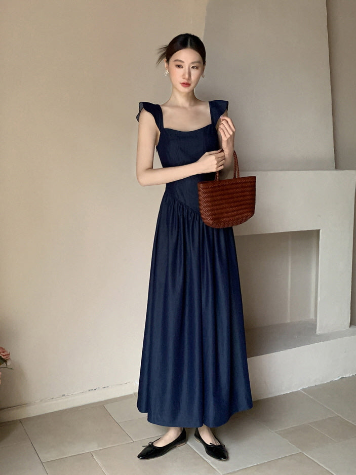 Wearing a navy vintage dress with flutter straps and a woven bag, the woman poses softly in a muted fairytale interior setting.