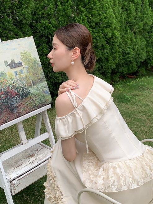 The model ties a ribbon shoulder strap on a cream ruffle top, seated beside a countryside painting in a romantic garden setting.