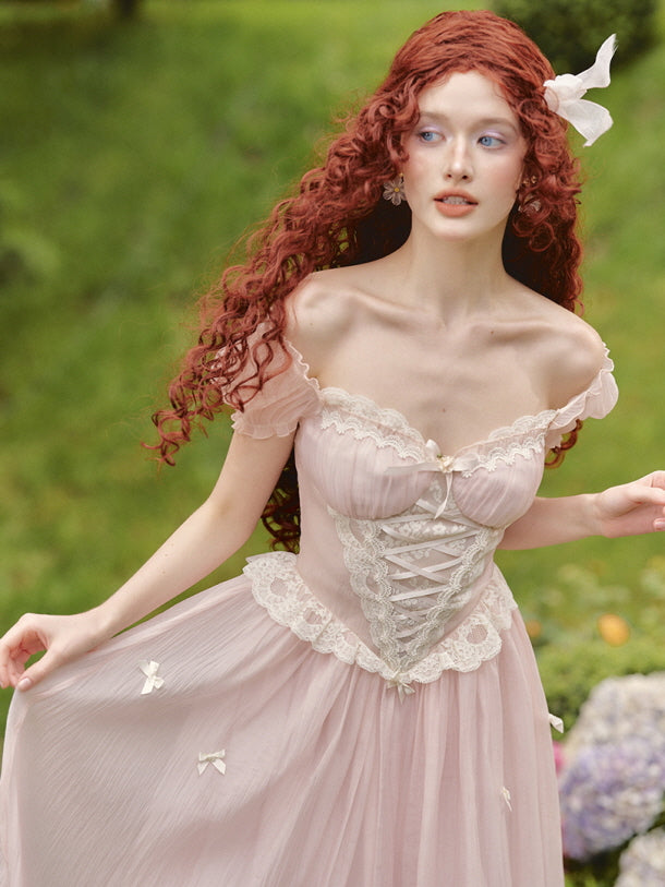 A model gently lifts the skirt of a blush pink corset fairytale dress with lace trim and ribbon bows, capturing a fairytale moment in a garden setting.