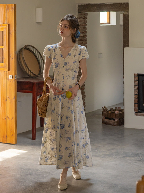 A woman wearing a cream floral maxi dress with blue flowers stands in a rustic, sunlit room holding a lemon. The dress features a v-neckline, button-down front, and cap sleeves, exuding a vintage cottagecore aesthetic.