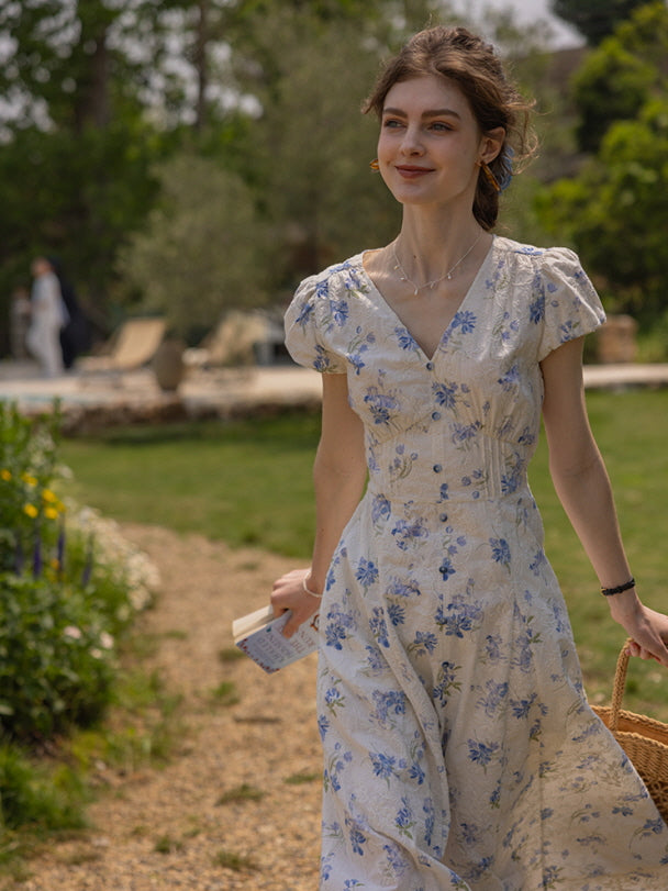 A woman walks outdoors in a vintage-inspired cream dress adorned with blue floral patterns, holding a book and a woven bag, exuding a soft cottagecore aesthetic.