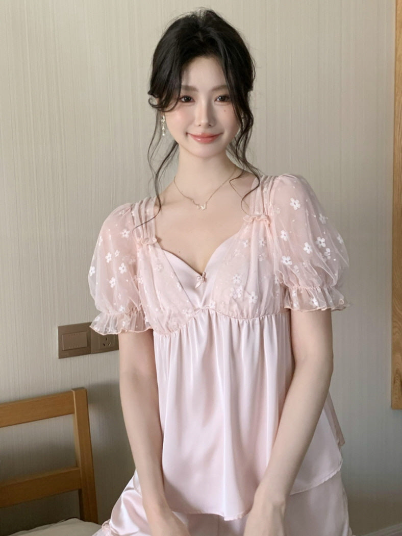 A woman smiles softly in a pink coquette loungewear set with floral mesh puff sleeves and a satin layered top, standing in a softly lit bedroom.
