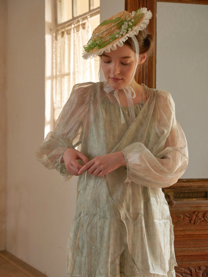 A woman in a sheer long-sleeve overlay gently fastens her dress ribbon, styled in a softly glowing vintage room with a fairytale mood.