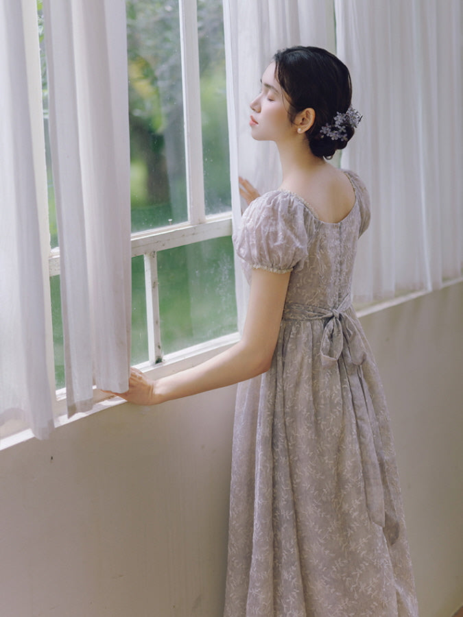 With her eyes closed and a flower hairpiece, the woman stands by the window in a lavender princesscore dress with a tied ribbon back.
