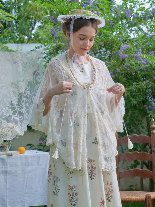 Standing in a floral dress and lace robe, the model gently ties the ribbon while surrounded by violet blossoms and vintage garden decor.