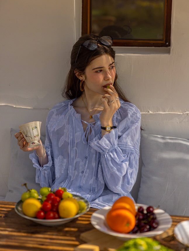 A young woman seated outdoors, enjoying fresh fruit while sipping tea from a floral teacup. She wears a blue floral blouse with delicate ruffle details, creating a dreamy, vintage-inspired aesthetic.