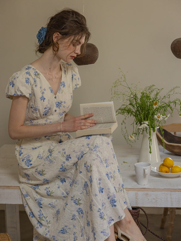A woman in a vintage-inspired floral blue and ivory dress sits on a rustic wooden table, reading a book. The dress features a delicate floral pattern and a V-neckline with button details, evoking a cottagecore aesthetic.
