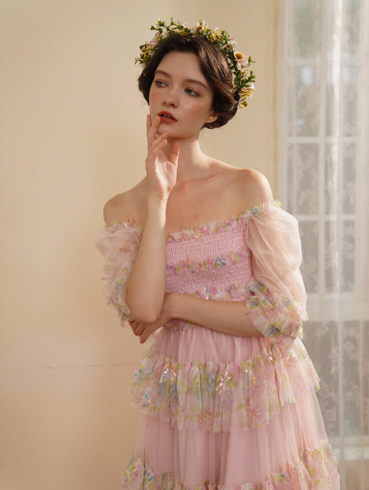 Wearing a pastel pink princesscore dress and floral crown, the model touches her lips thoughtfully in soft natural light by the window.