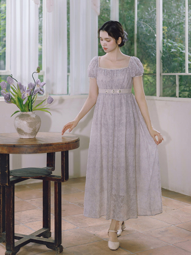 A woman walks gracefully beside a wooden table in a lavender puff-sleeve dress with floral lace waist, capturing a dreamy vintage fairytale look.