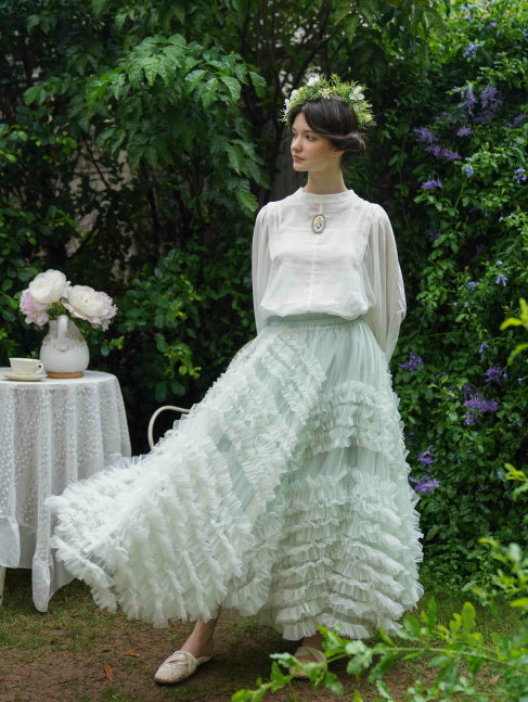 A model in a white embroidered blouse and pastel green ruffled skirt poses gracefully in a garden, embodying a romantic fairycore aesthetic.