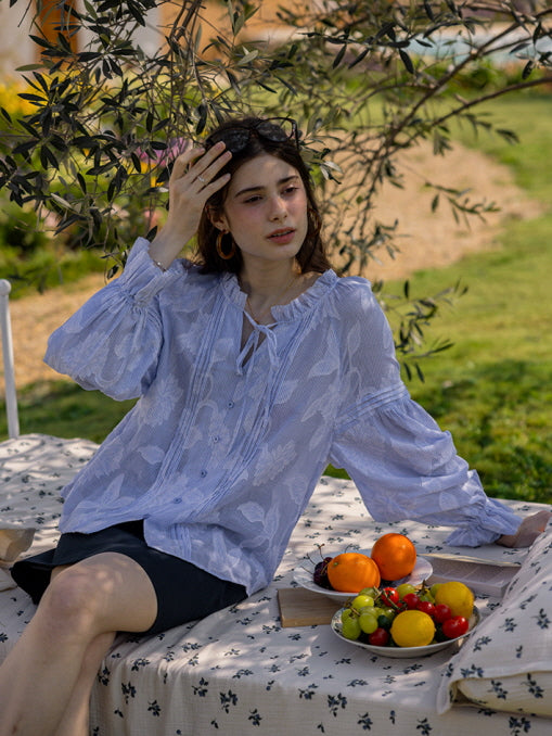 A young woman relaxing outdoors under a tree, wearing a blue floral blouse with puffed sleeves and delicate ruffle details. A tray of fresh fruit adds a charming, fairytale-inspired touch to the serene scene.