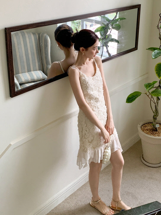 Leaning against a mirror-lit wall, she wears a vintage-inspired ivory dress with floral textures and chiffon hem, holding a pearl bag in hand.