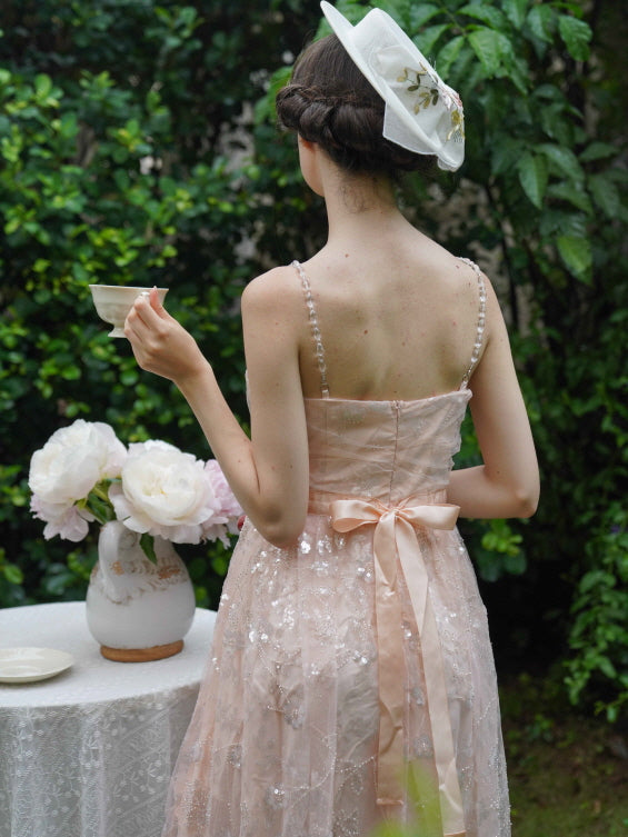 Back view of a model holding a teacup in a blush sequin dress with ribbon waist detail, evoking a refined vintage tea party in a garden.