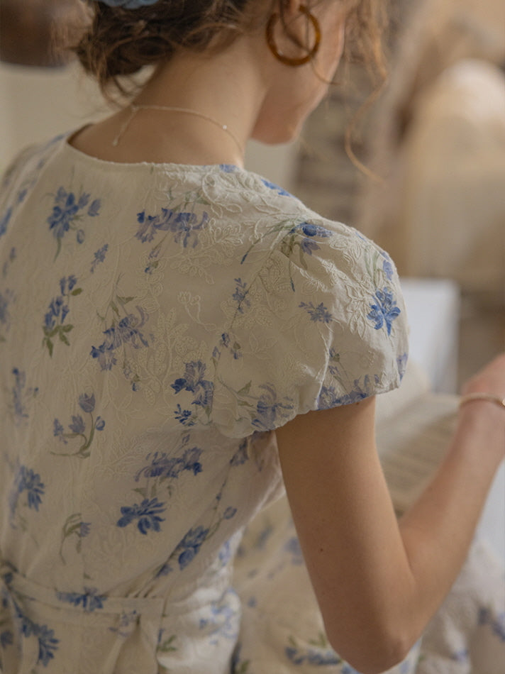 A close-up view of the back of a floral blue and ivory dress, showcasing delicate puff sleeves and a subtle waist tie. The textured fabric features a vintage-inspired floral pattern, exuding a soft, romantic aesthetic.