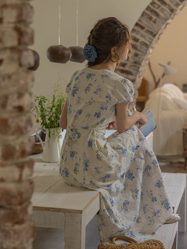 A woman in a blue floral vintage dress sits on a rustic wooden table, reading a book with a calm expression. The soft, cottagecore ambiance is complemented by hanging pottery decor and a bouquet of wildflowers.