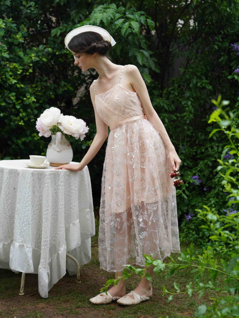 Model leans over a lace-covered tea table in a blush pink embroidered dress with pearl straps, capturing a soft princesscore garden moment.