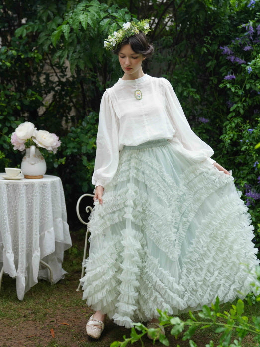 The model lifts her pastel ruffle skirt gently while wearing a romantic blouse, embodying soft fairycore charm in an enchanted outdoor setting.
