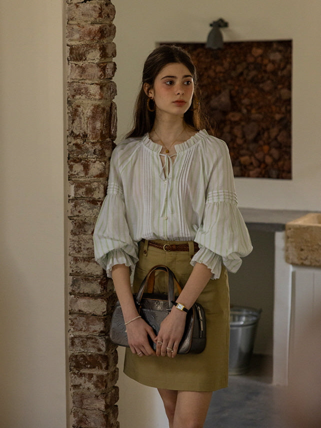 A young woman leaning against a brick wall, carrying a gray handbag while wearing a light green and white striped blouse with puffed sleeves and a khaki skirt. The warm, vintage aesthetic creates a refined, fairytale-inspired look.