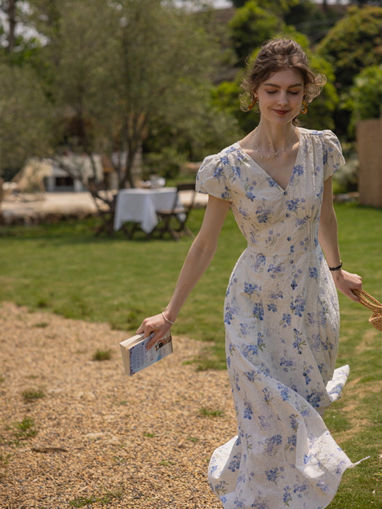 A serene outdoor scene where a model walks along a gravel path in a blue floral dress, holding a book and a woven bag, capturing a dreamy, cottagecore aesthetic.