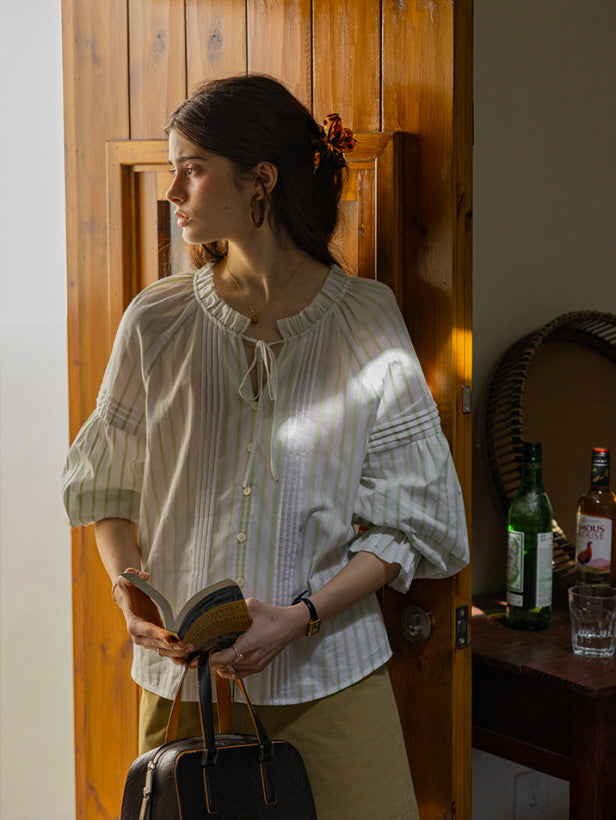 A young woman standing by a wooden door, holding a book and a black handbag while wearing a light green and white striped blouse with ruffled cuffs and a tie neckline. The soft, vintage-inspired look exudes a relaxed, cottagecore vibe.