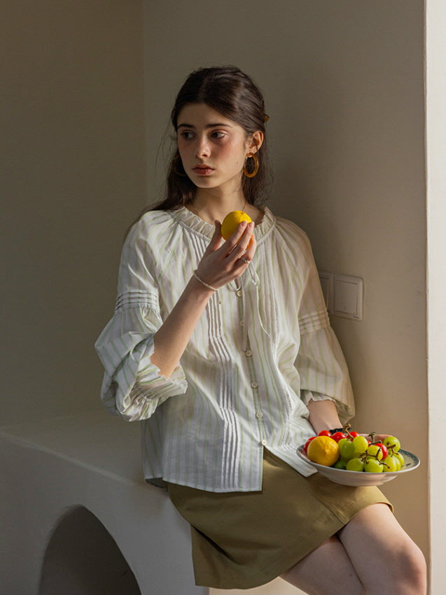 A young woman seated by a sunlit wall, holding a lemon and a plate of fresh fruit. She wears a light green and white striped blouse with puffed sleeves and a tie neckline, creating a soft, cottagecore-inspired look.